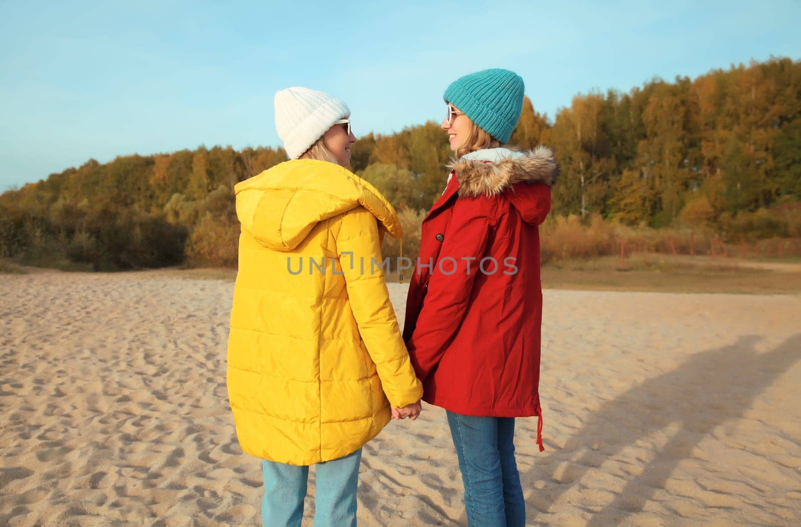 Happy young two women friends together on sunny beach, stylish smiling girlfriends standing on sea coast in glasses, colorful red yellow coat jacket and hat outdoors