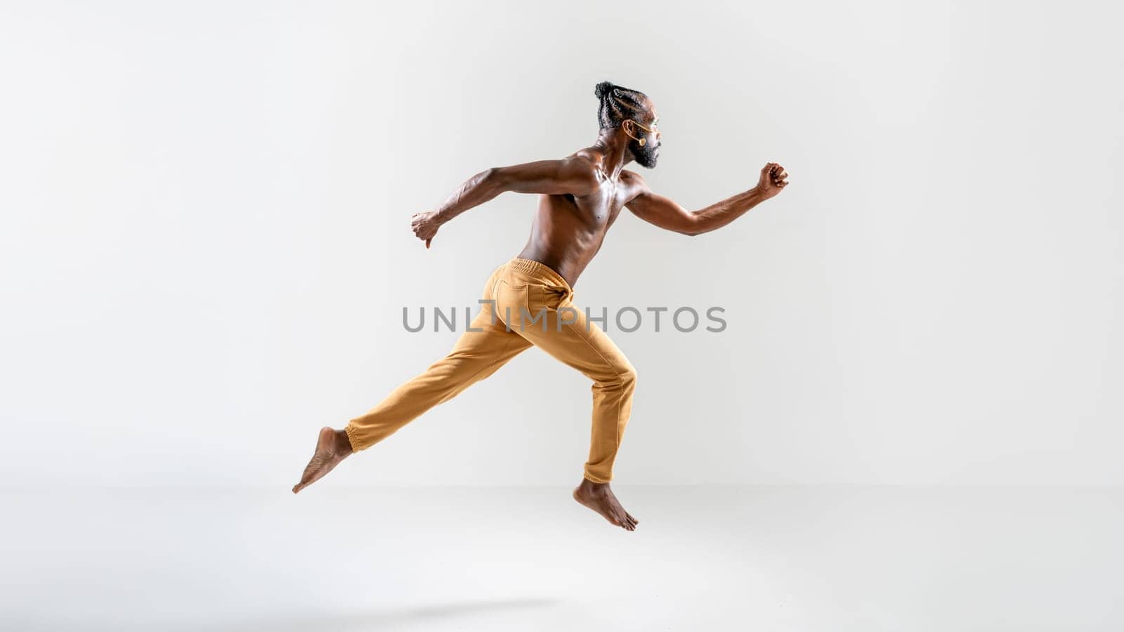 Determined shirtless gay model jumping over gray background. Full body side view of strong young man is in midair at plain studio.