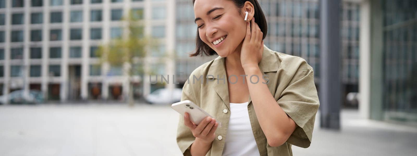 Smiling asian girl listens music in wireless headphones, looks at her phone, choosing music or podcast. Young woman calling someone, using headphones.