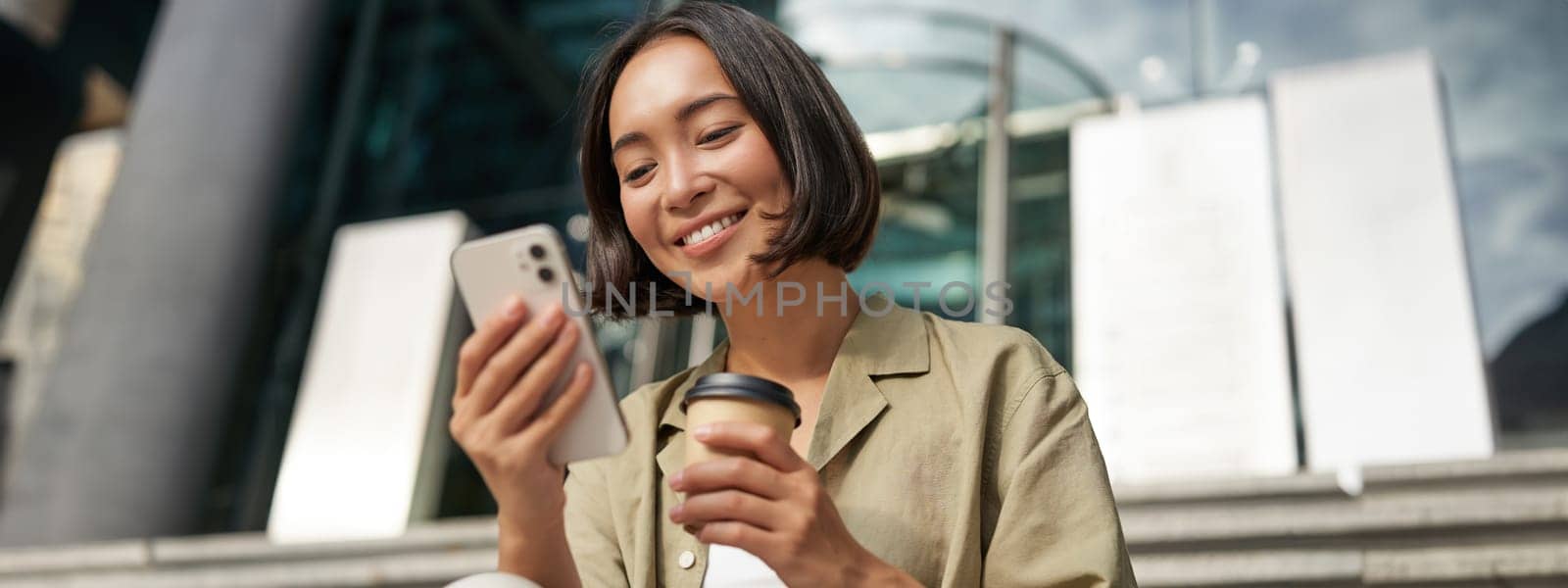 Beautiful smiling asian girl, drinking coffee, using mobile phone and sits on stairs outside. Young woman video chat outdoors by Benzoix