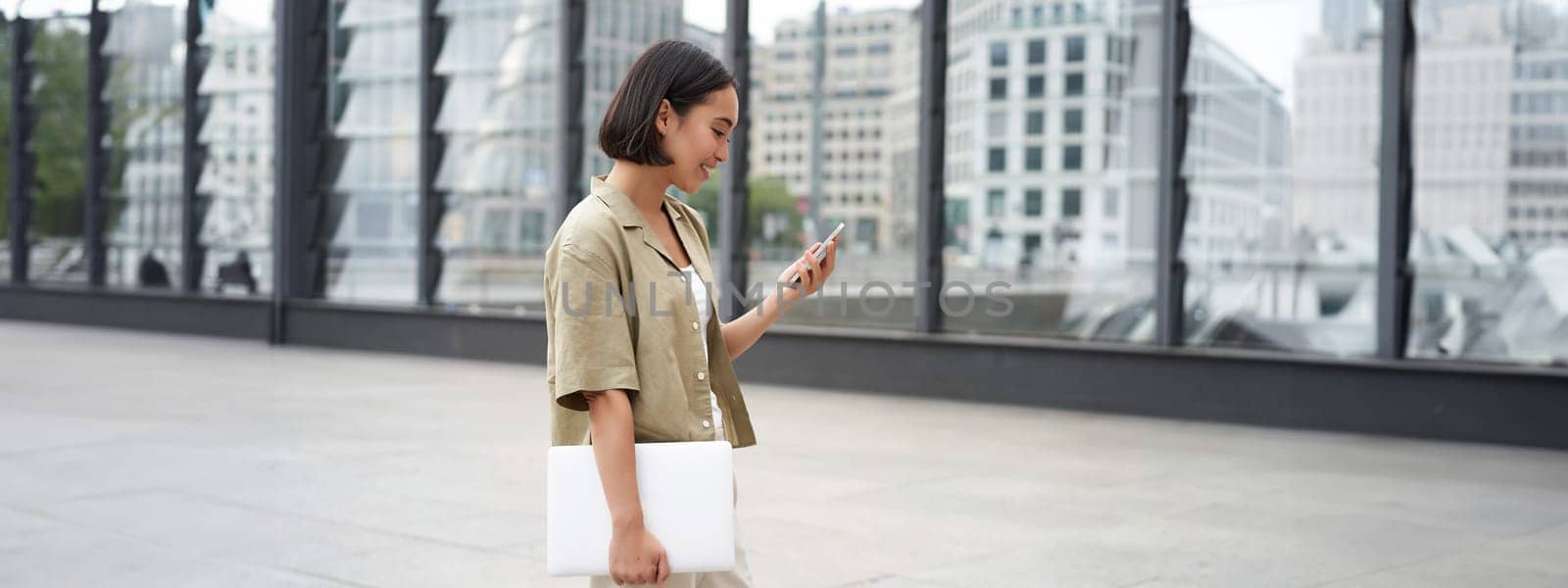 Young asian woman with laptop, walking on street and texting message, smiling while looking at smartphone.