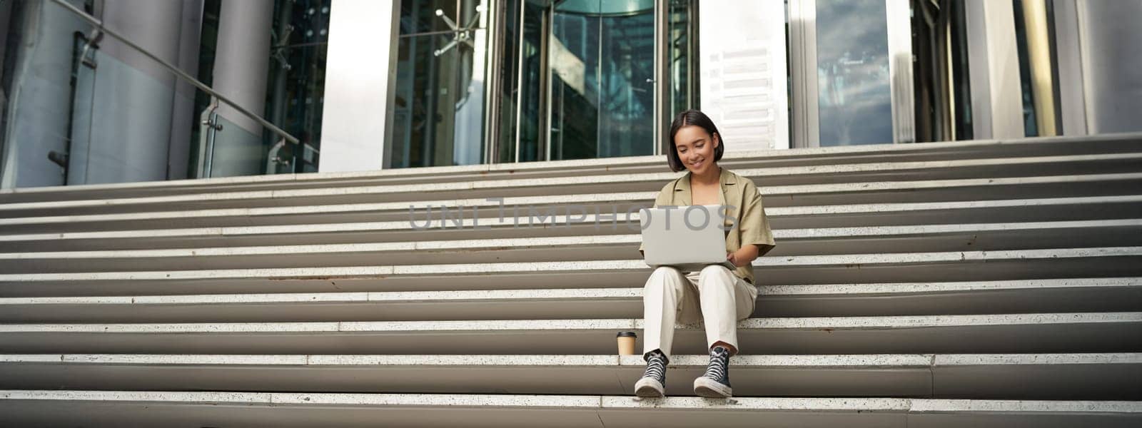 Asian girl student sits on stairs near campus, types on laptop, does her homework outdoors.