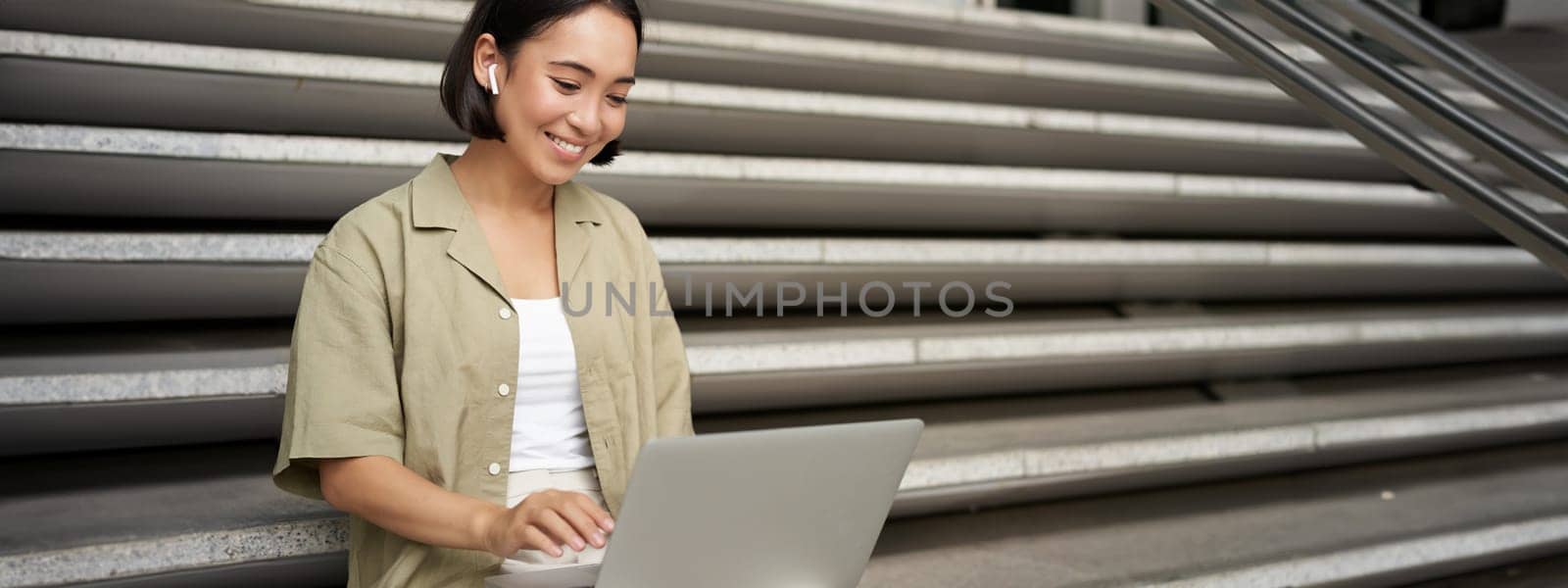 Remote worker. Smiling asian girl sits outdoors on street with laptop. Happy young woman working on computer remotely, listen music in wireless headphones.