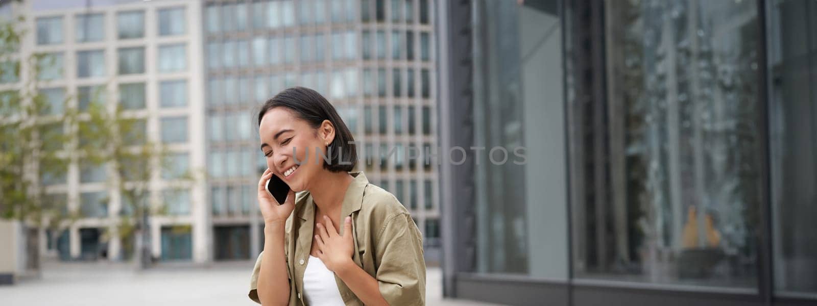 Vertical shot of smiling stylish asian girl, having a telephone conversation and walking on street. Young woman talking on mobile phone.