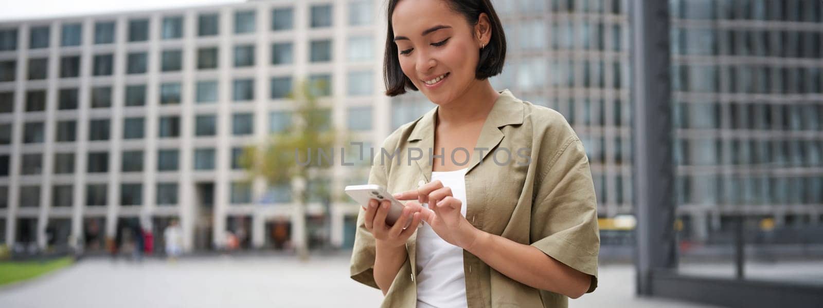 Mobile technology. Smiling asian woman using smartphone app, looking at her telephone on street, checking map, calling or texting someone.