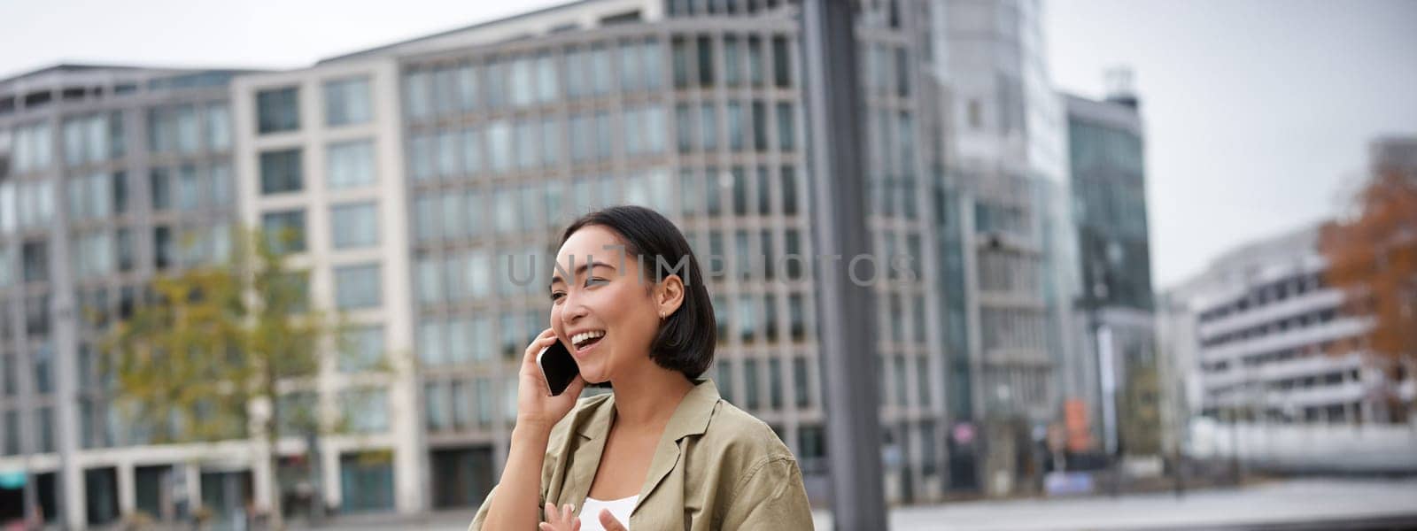Vertical shot of smiling stylish asian girl, having a telephone conversation and walking on street. Young woman talking on mobile phone.