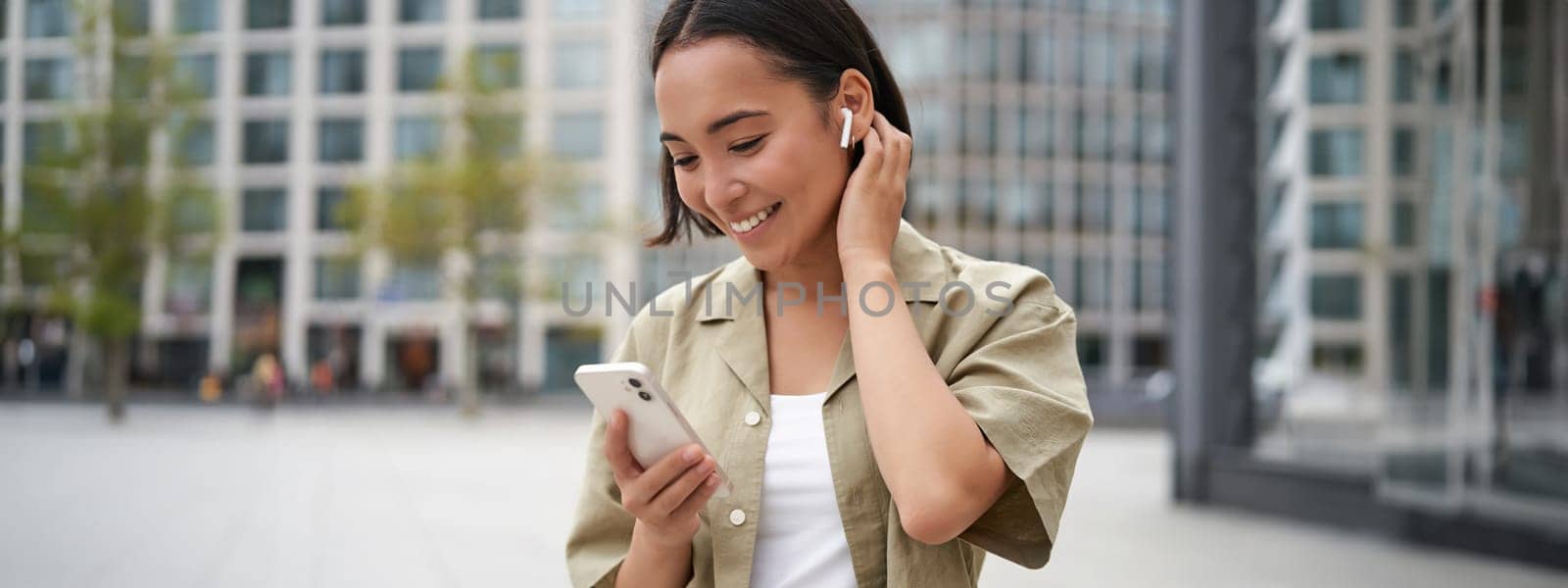 Smiling asian girl listens music in wireless headphones, looks at her phone, choosing music or podcast. Young woman calling someone, using headphones by Benzoix