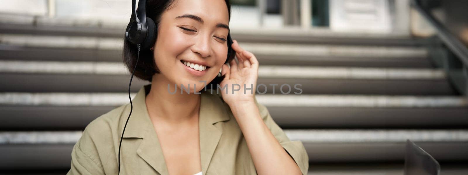 Happy asian girl listens to music in headphones, smiles, sits outdoors on stairs by Benzoix