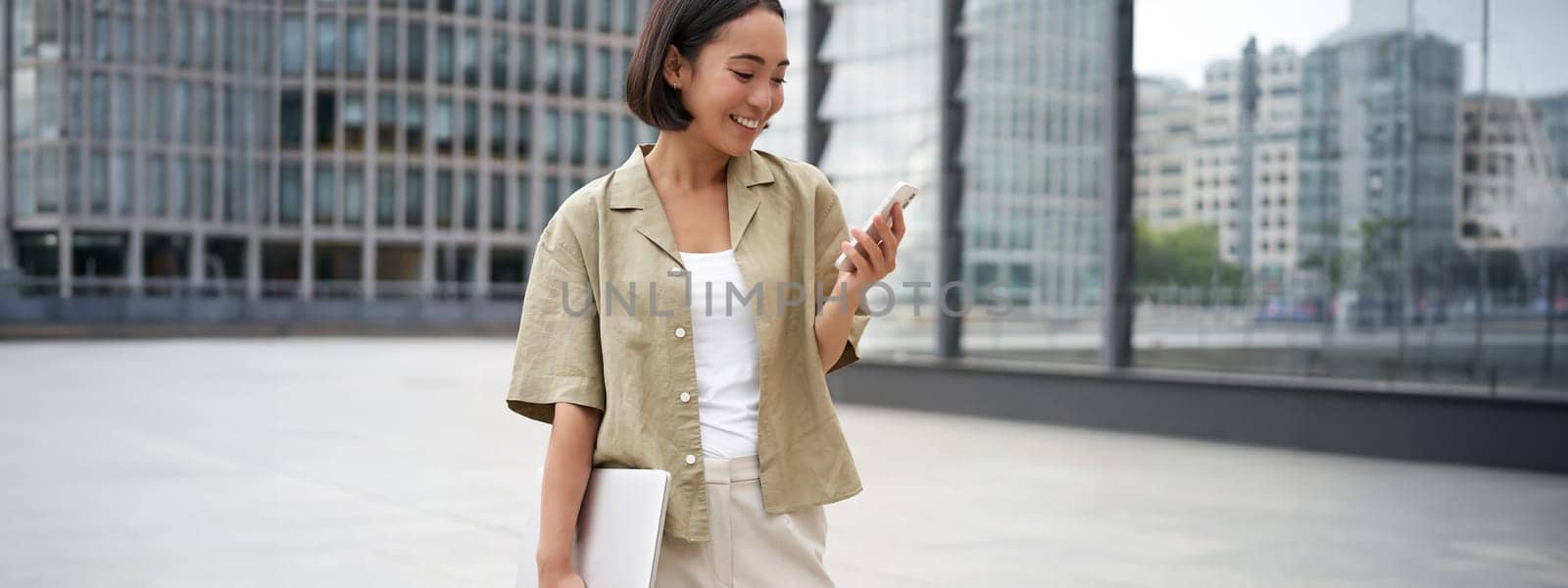 Young asian woman with laptop, walking on street and texting message, smiling while looking at smartphone.