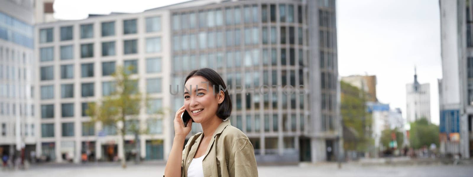 Modern young asian girl talks on mobile phone, uses telephone on city street. Woman smiling while calling someone on smartphone by Benzoix