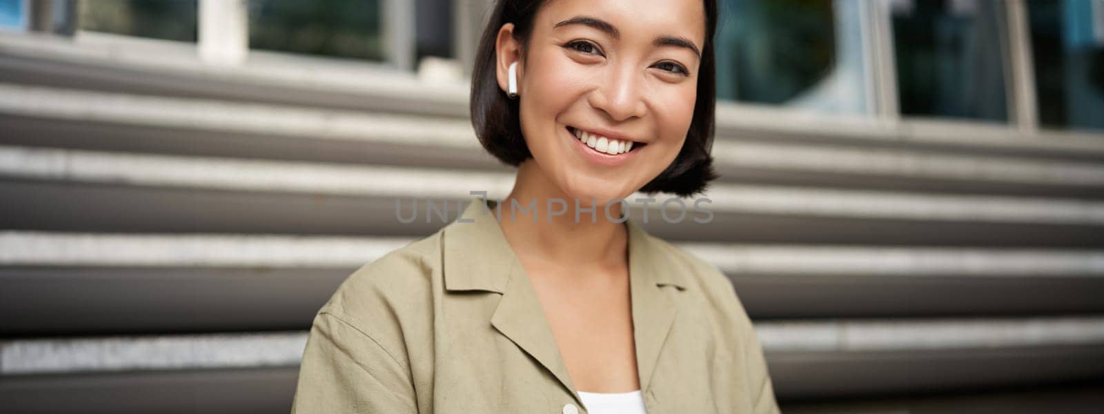 Portrait of smiling asian girl listens music, podast in wireless earphones, using headphones outdoors, sitting on street by Benzoix