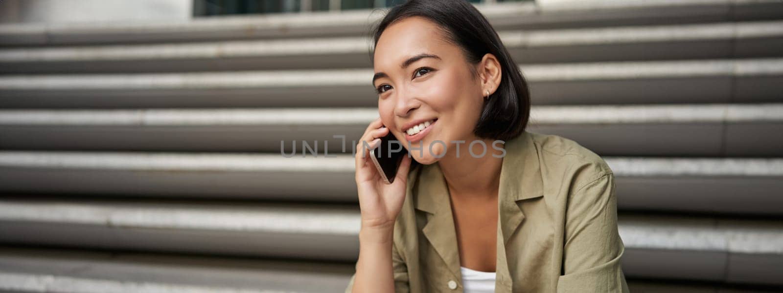 Portrait of beautiful asian girl talks on mobile phone, sits on street stairs. Woman with smartphone smiling, making a call.
