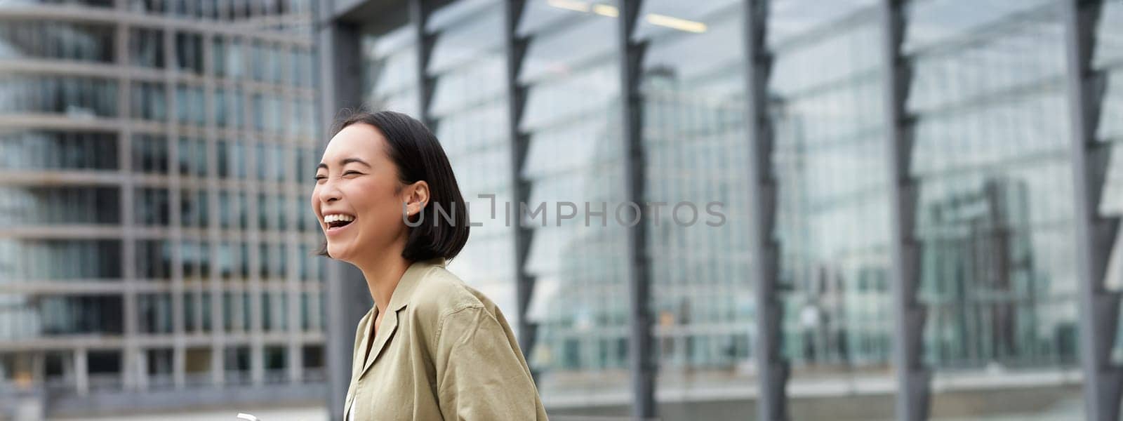 Vertical shot of beautiful asian woman walking on street, laughing and looking happy, enjoying the day by Benzoix