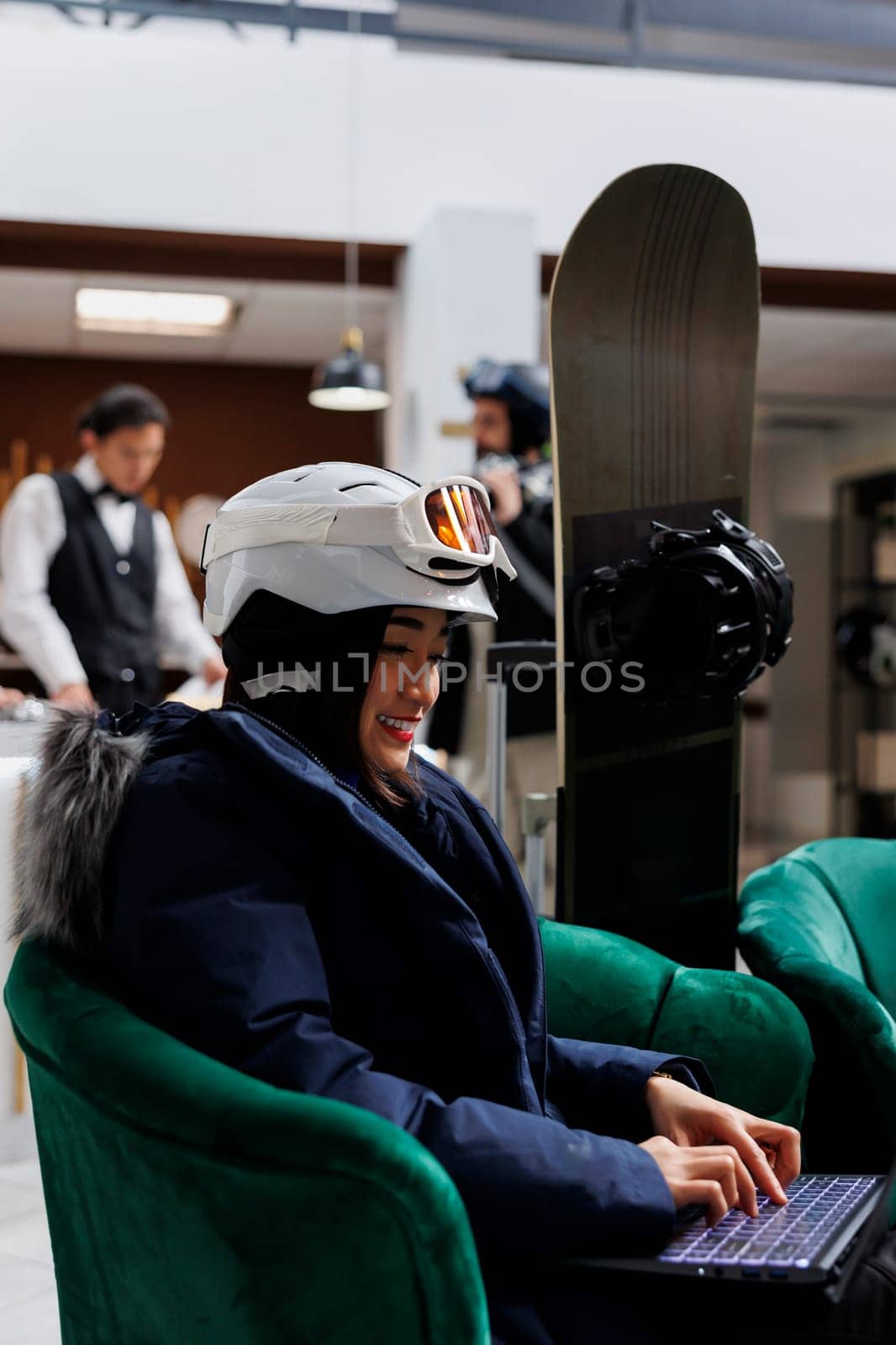 Detailed view of woman seated in hotel lobby perusing the web on laptop. Close-up of female guest clothed in winter gear and with snowboarding equipment nearby uses digital device in ski resort.