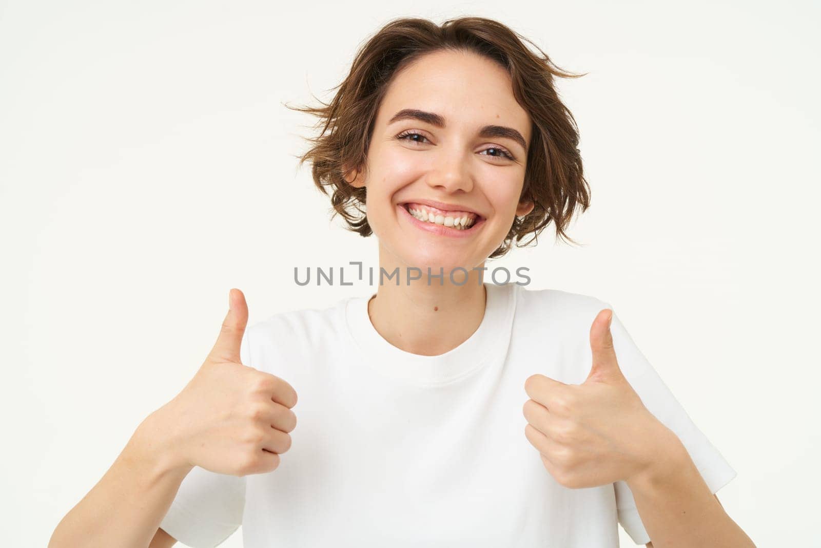 Close up of happy brunette woman, shows thumbs up, approves something, recommends, gives positive feedback, stands over white background.