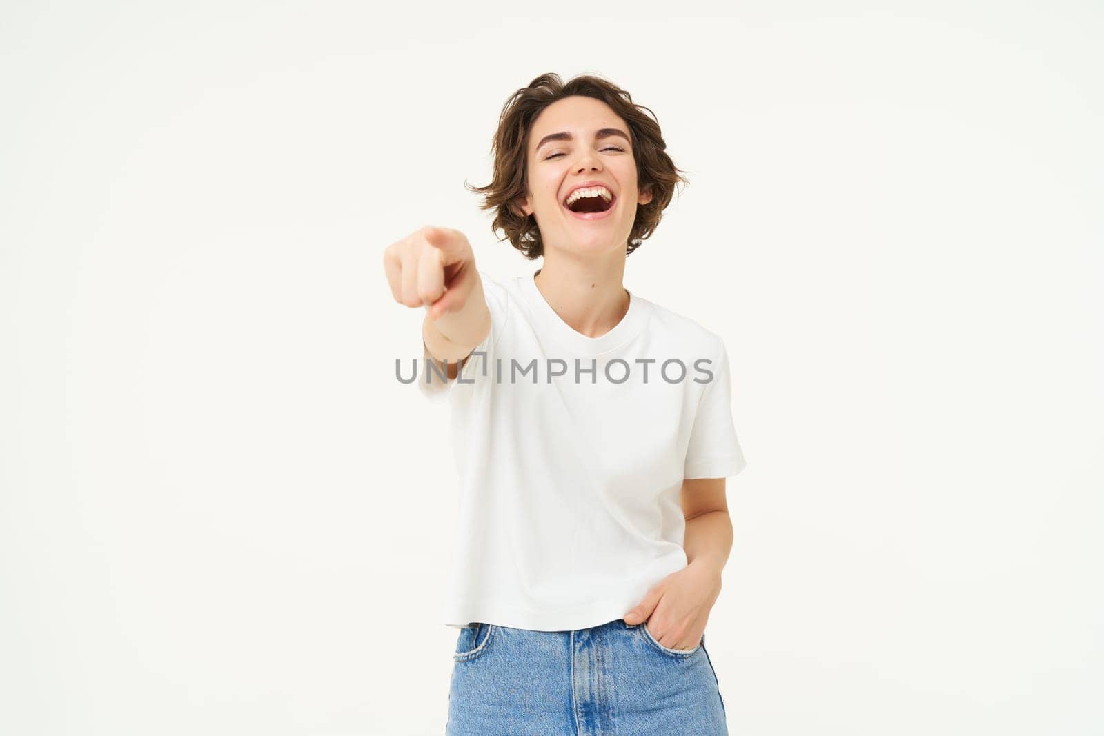 Portrait of enthusiastic woman laughing, pointing finger at camera and smiling, choosing you, inviting people, standing over white background. Copy space
