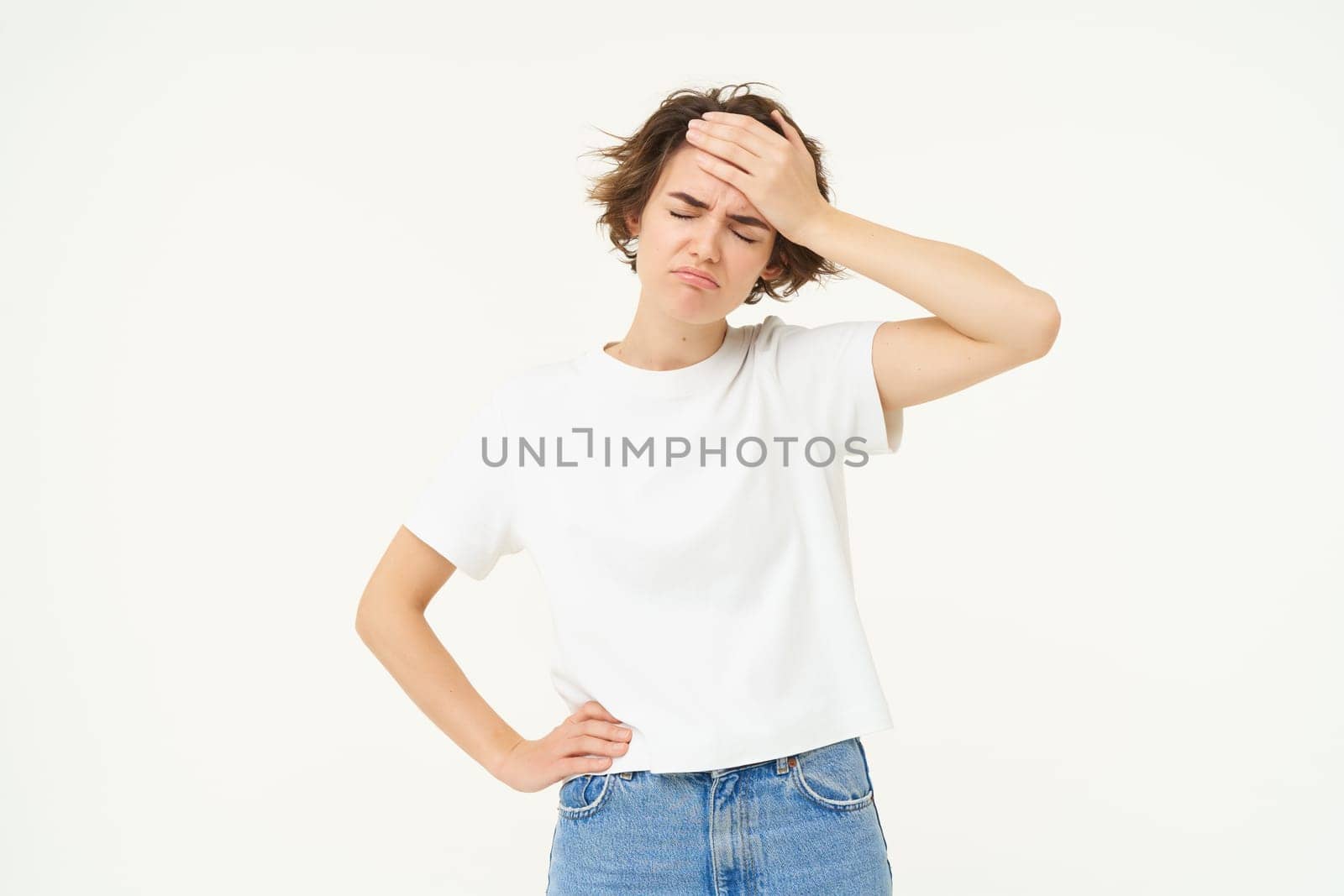 Health and people. Young woman has headache, touches her head and grimacing from painful migraine, feeling unwell, dizzy, standing over white background.