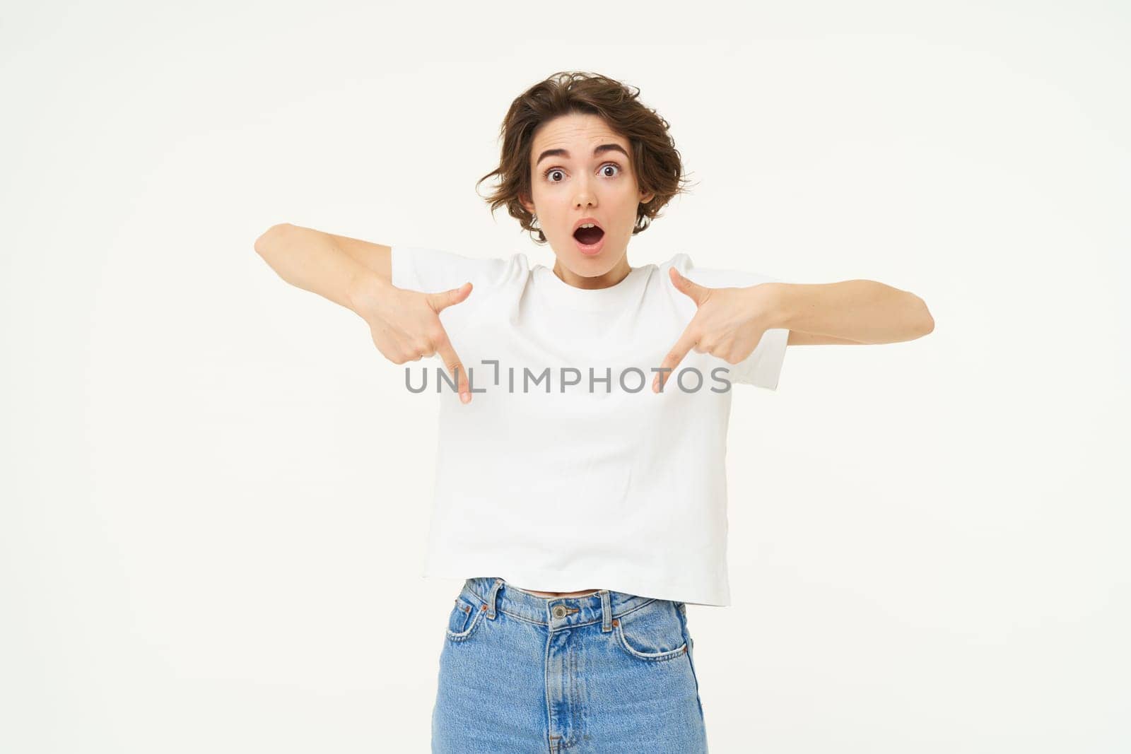 Portrait of amazed girl, pointing fingers down, showing something and looking surprised, standing over white studio background.
