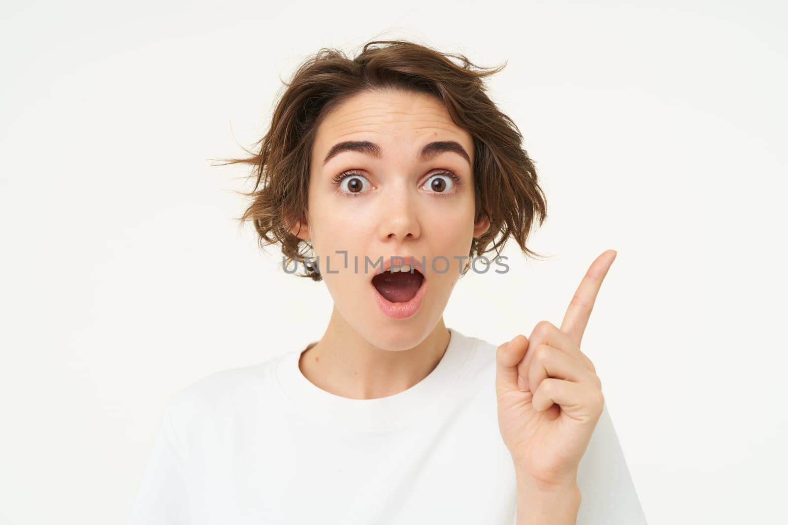 Excited girl has an idea, thinks of solution, pointing one finger and smiling, standing over white background.