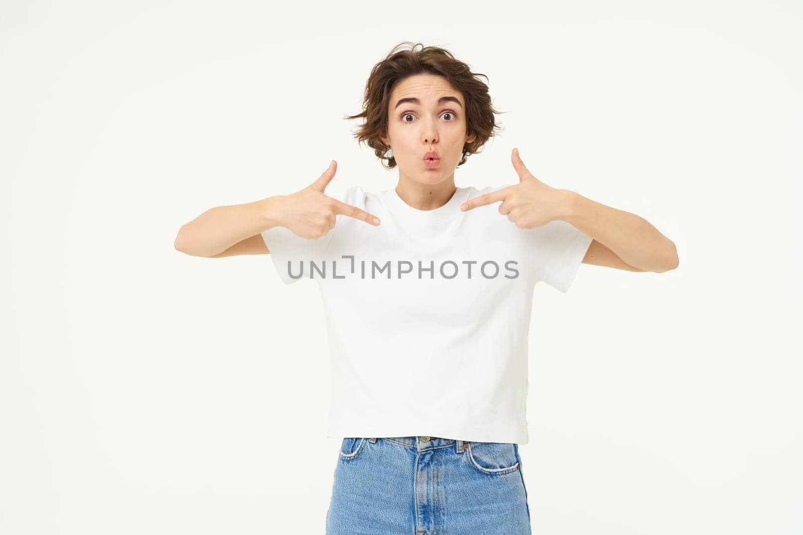 Image of girl with surprised look, pointing at herself with amazed face, being chosen or winning something, standing over white background.