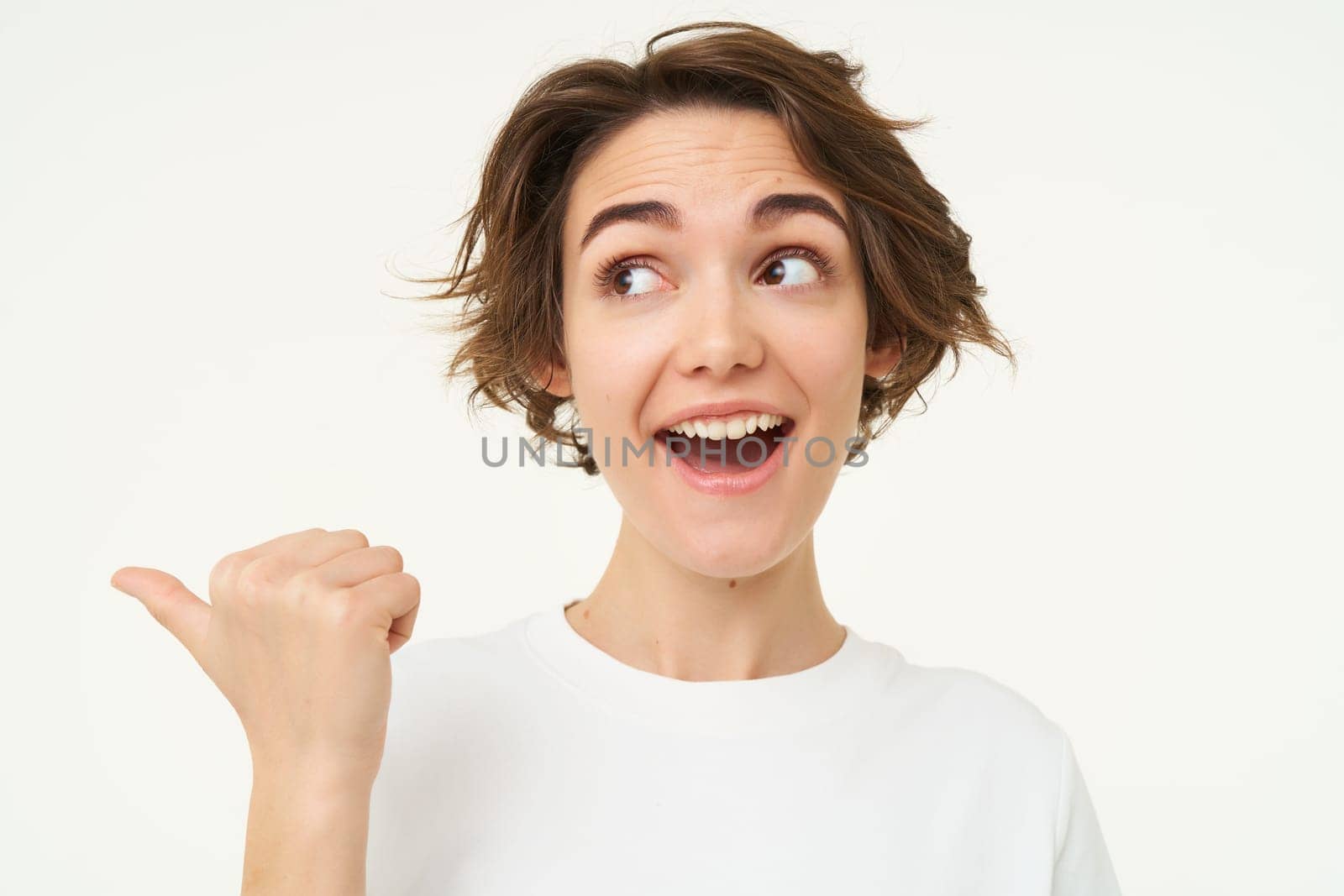 Image of excited brunette woman with short haircut, pointing left, smiling and looking happy, showing you advertisement, standing over white background by Benzoix