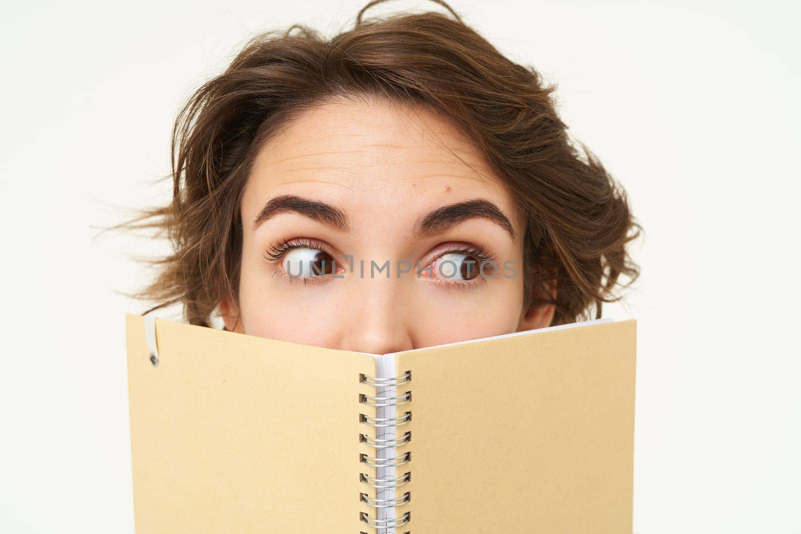 Close up portrait, young woman hiding her face behind planner, holding notebook and looking sideways, side eye, standing over white background by Benzoix