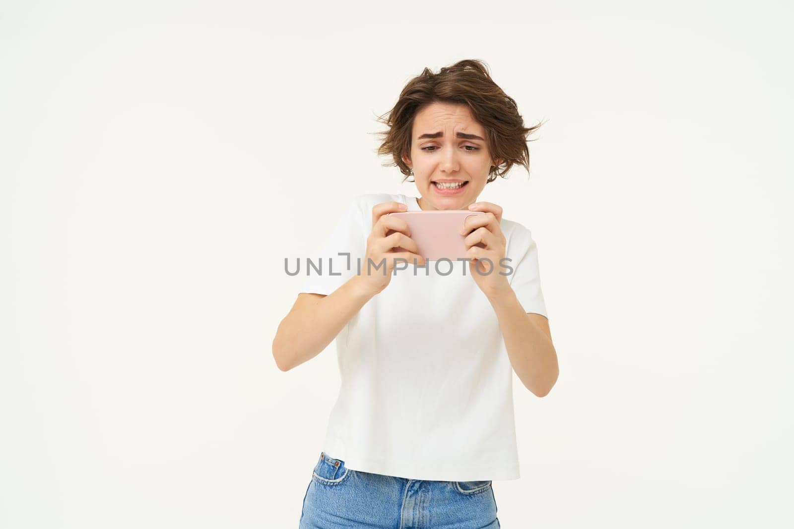 Portrait of woman playing video games on mobile phone, holding smartphone with both hands, standing over white studio background.