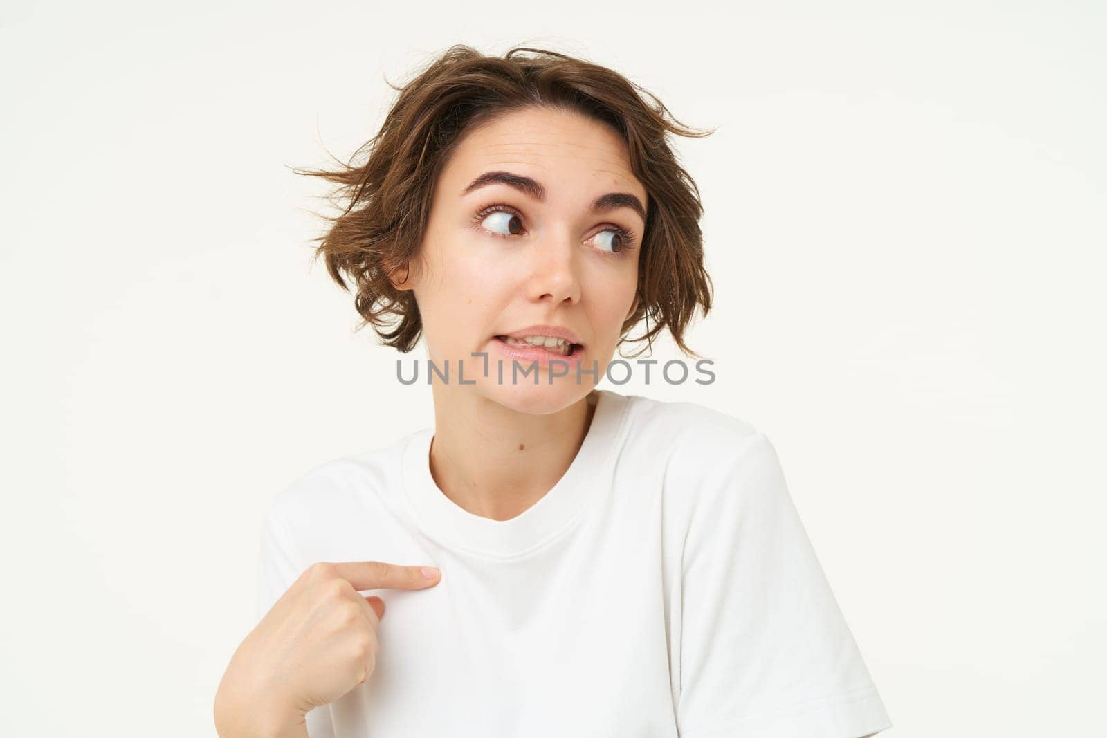 Portrait of awkward young woman, oops face, looks guilty, points at herself, being blamed or accused, stands over white background.