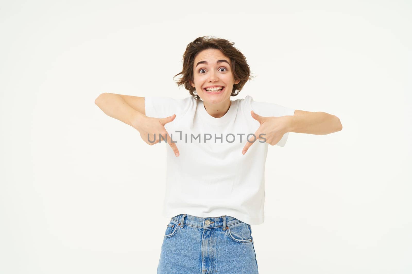 Portrait of cute, happy young woman pointing fingers down, showing advertisement, demonstrating banner on the bottom, standing against white background by Benzoix