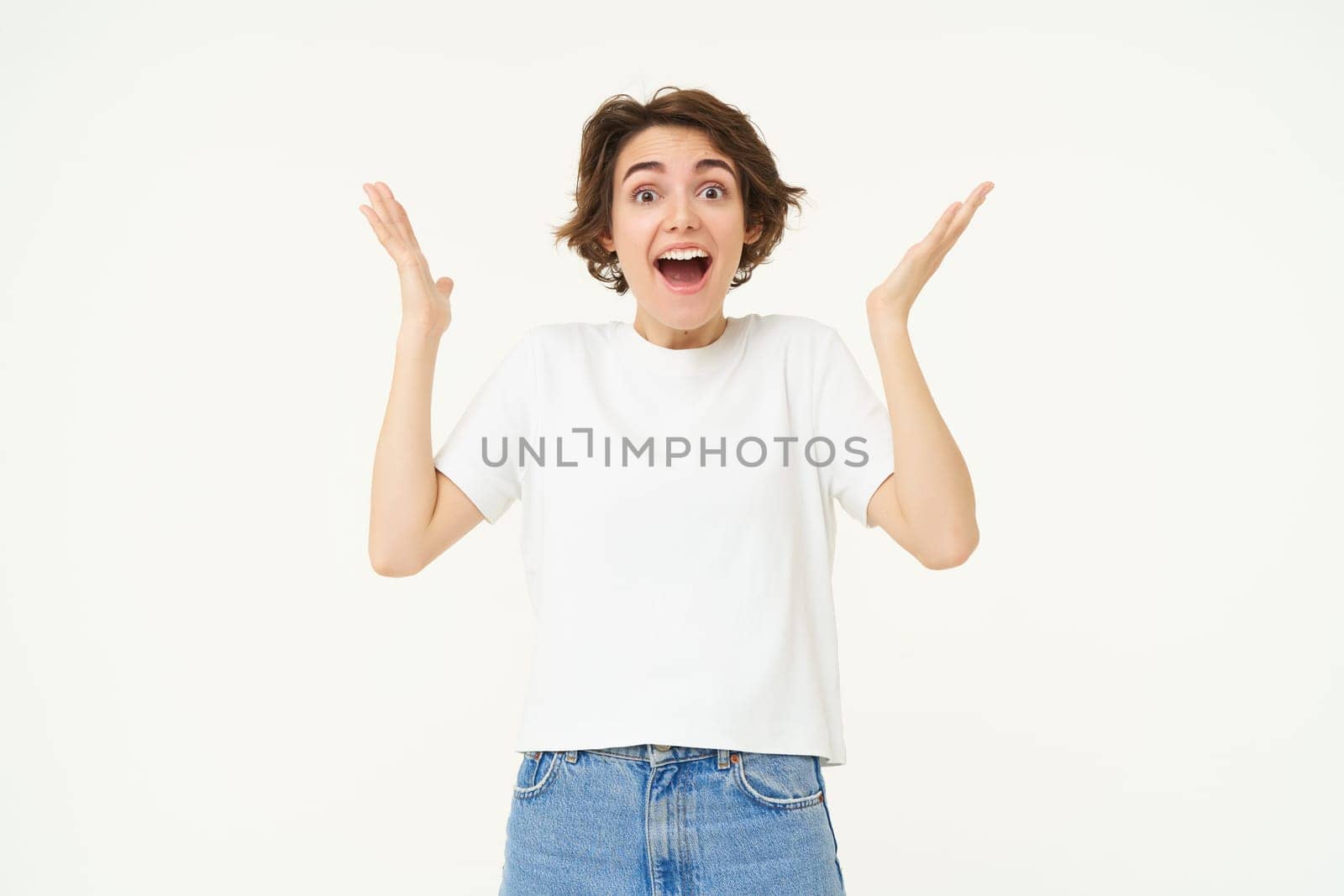 Portrait of amazed, happy young woman, raising hands up and looks surprised, standing excited against white background.
