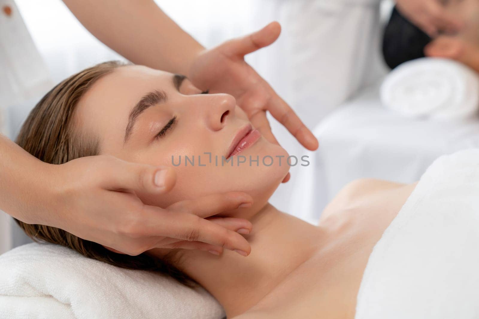 Caucasian couple enjoying relaxing anti-stress head massage and pampering facial beauty skin recreation leisure in dayspa modern light ambient at luxury resort or hotel spa salon. Quiescent