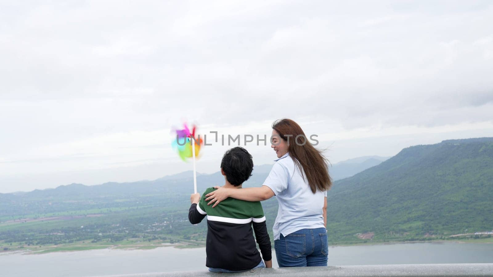 A progressive woman and her son are on vacation, enjoying the natural beauty of a lake at the bottom of a hill while the boy carries a toy windmill.