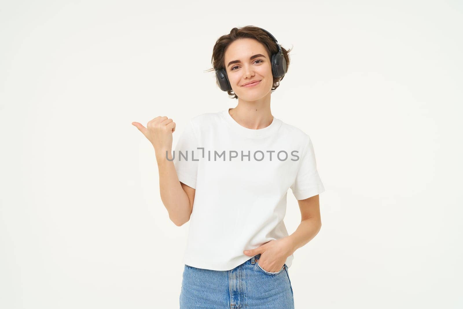 Portrait of brunette woman, smiling with confidence, pointing finger left, showing advertisement, standing over white background.