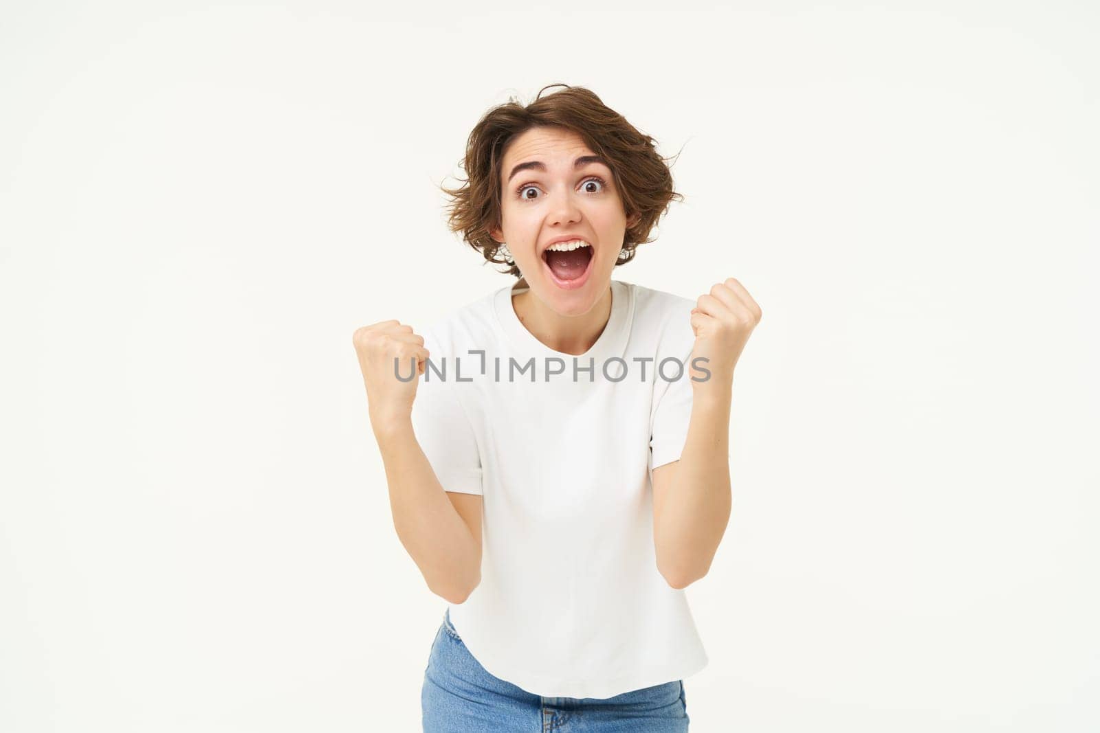 Cheerful brunette woman winning, triumphing, celebrating victory, achieves goal, stands over white background.