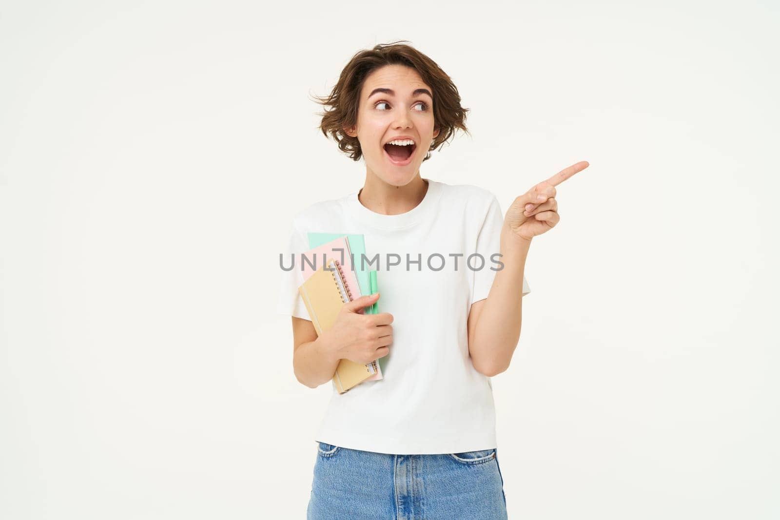 Portrait of woman, student with amazed face, carry her notebooks and documents, points at upper right corner, shows advertisement, studio background.