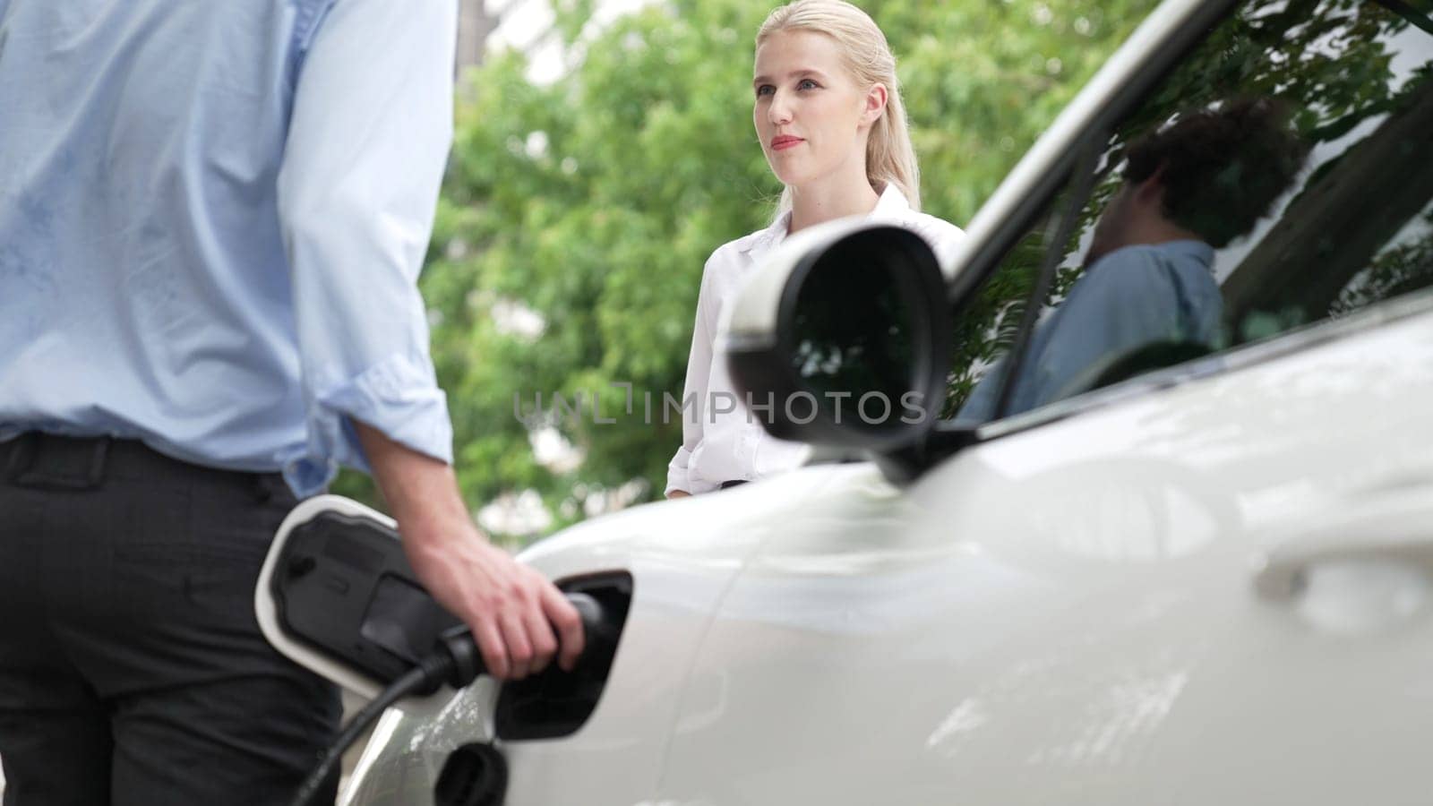 Progressive businessman and businesswoman install charger plug from charging station to electric car before driving around city center. Eco friendly rechargeable car powered by clean energy.