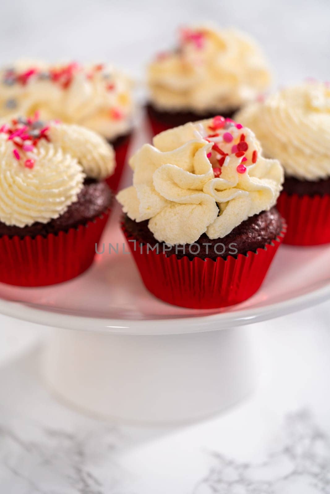 Freshly baked velvet cupcakes with white chocolate ganache frosting decorated with sprinkles.