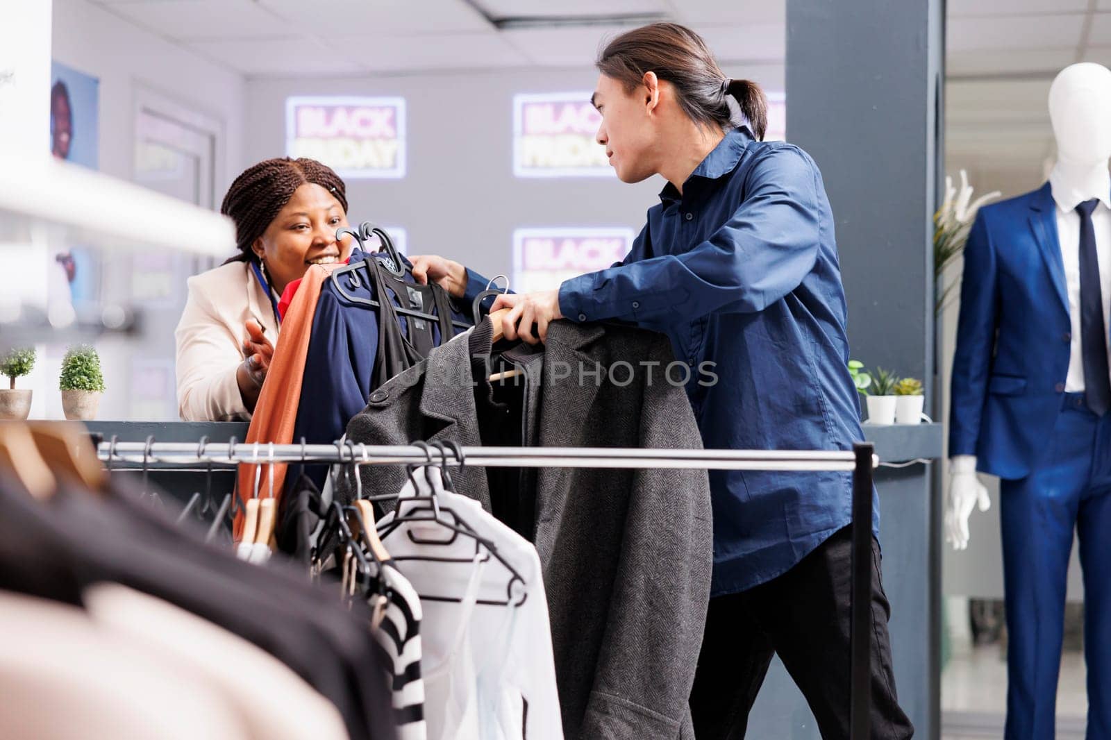 Friendly smiling African American woman retail worker helping male customer to choose clothes on Black Friday. Pleasant female cashier serving shopper in clothing store during sales season