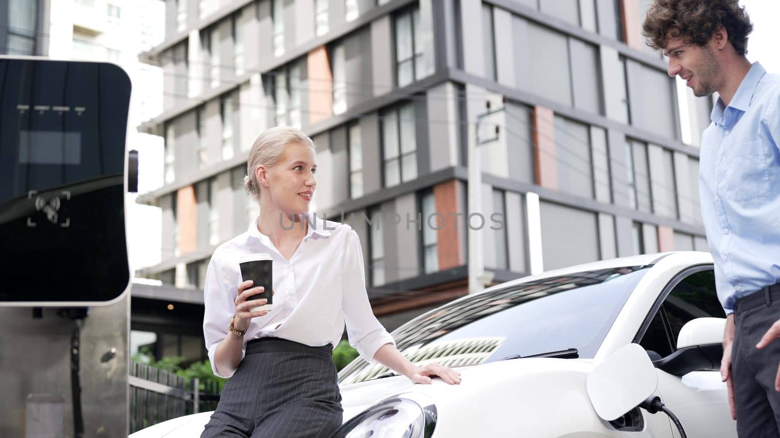 Progressive businessman and businesswoman leaning on electric car connected to charging station before driving around city center. Eco friendly rechargeable car powered by clean energy.