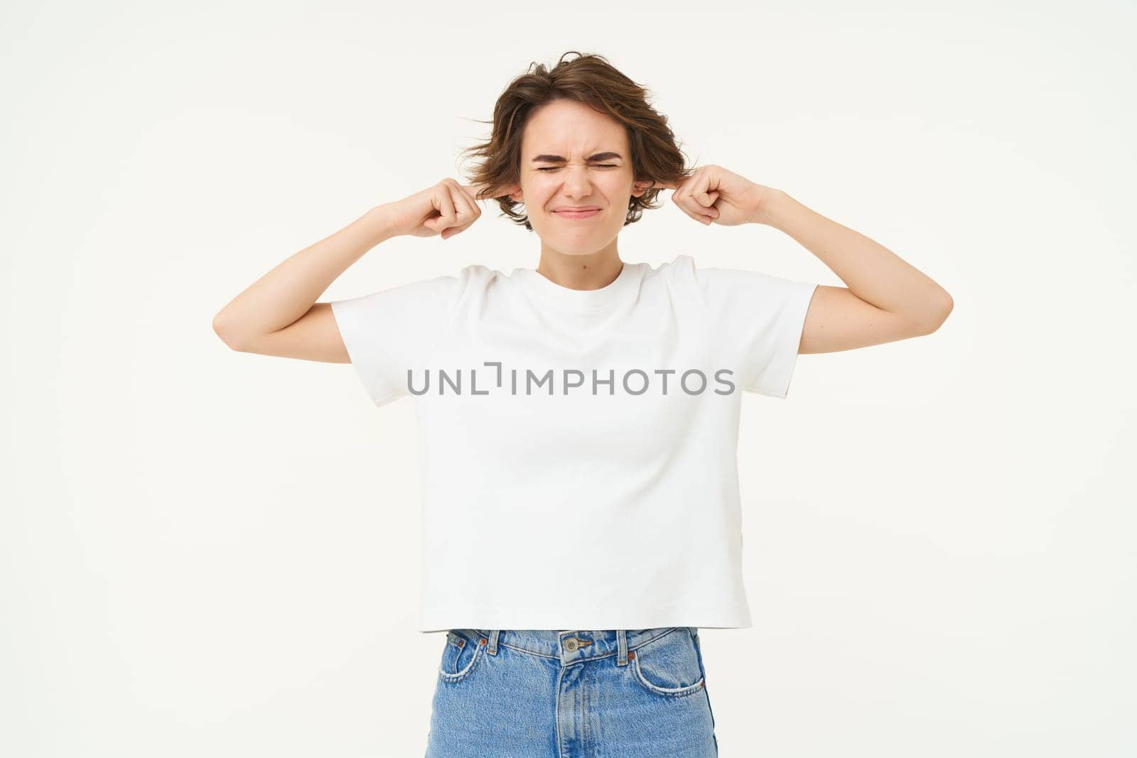 Woman standing annoyed, holding fingers in ears, unwilling to listen, feeling discomfort from loud noise, standing over white background by Benzoix