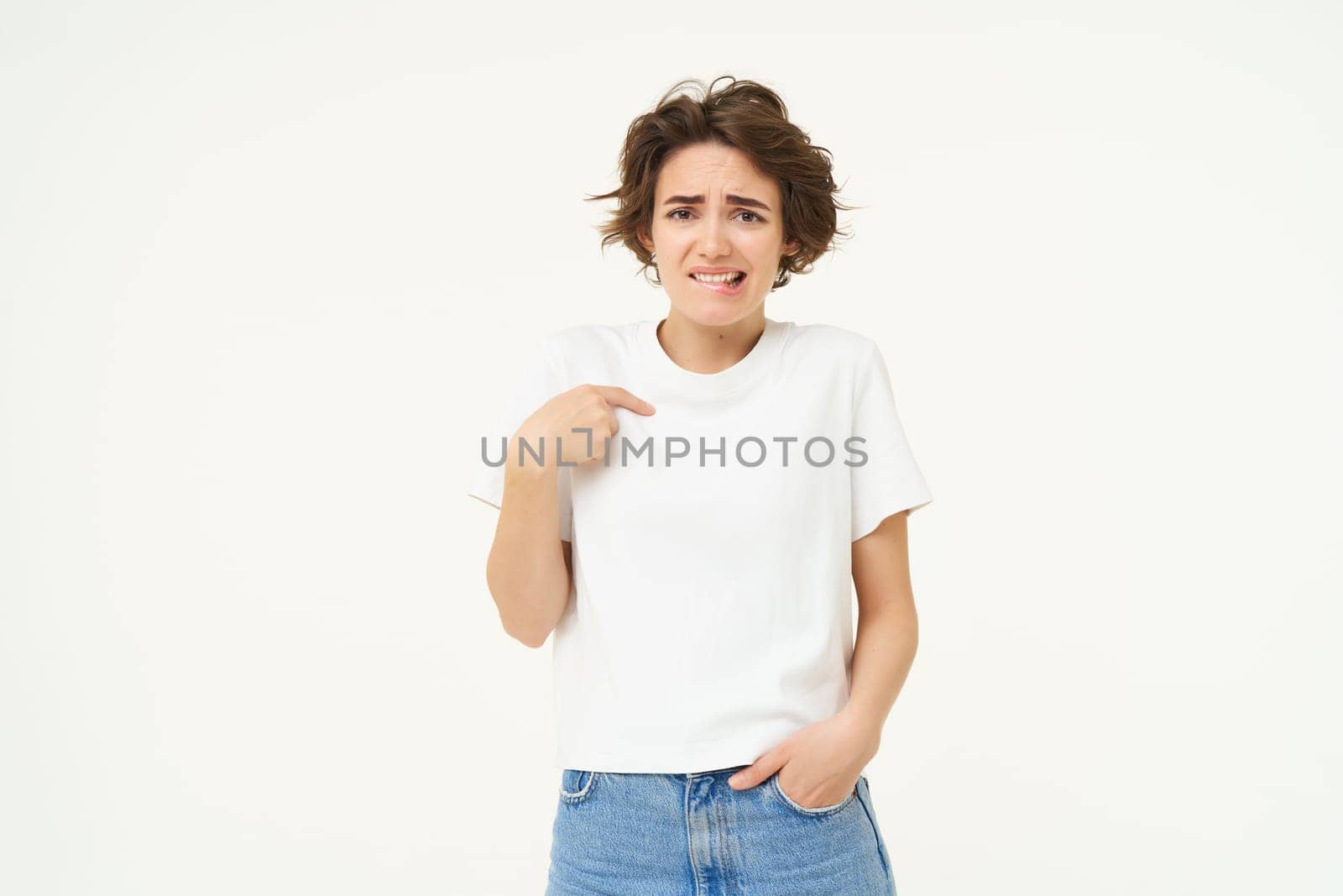 Portrait of awkward girl, feeling uncomfortable, pointing at herself with unsure face, posing over white background.