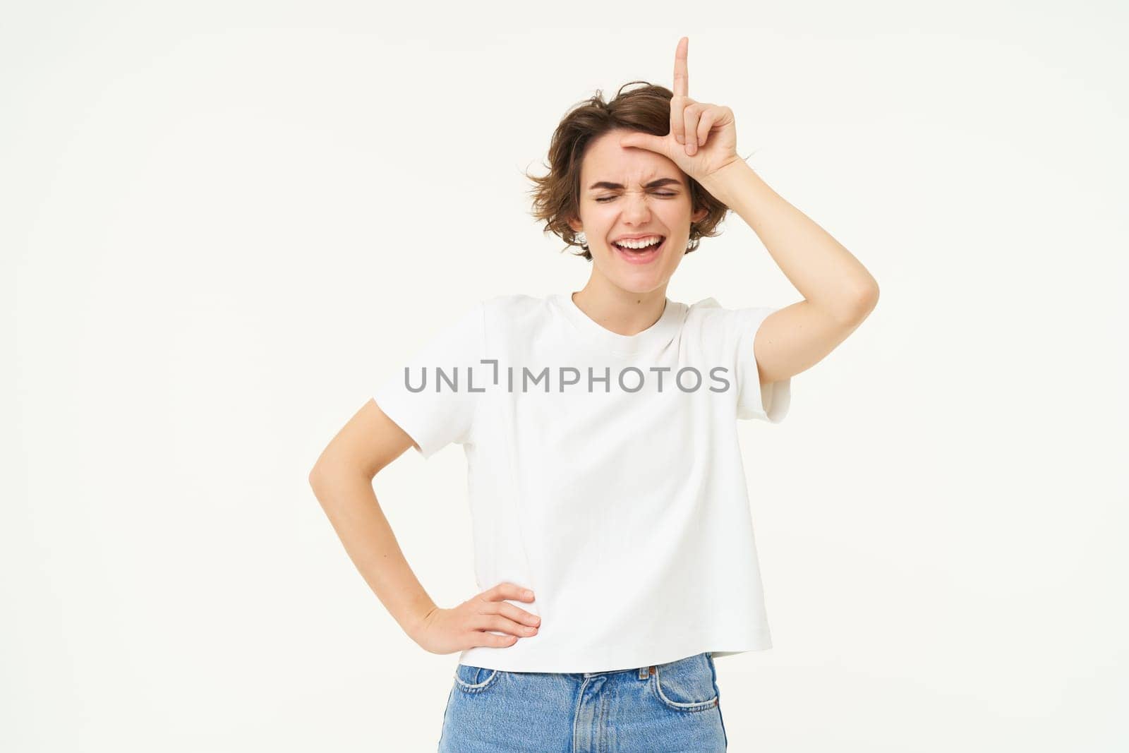 Portrait of happy young woman mocking, winning prize and making fun of loser, showing l letter on forehead, stands over white background.