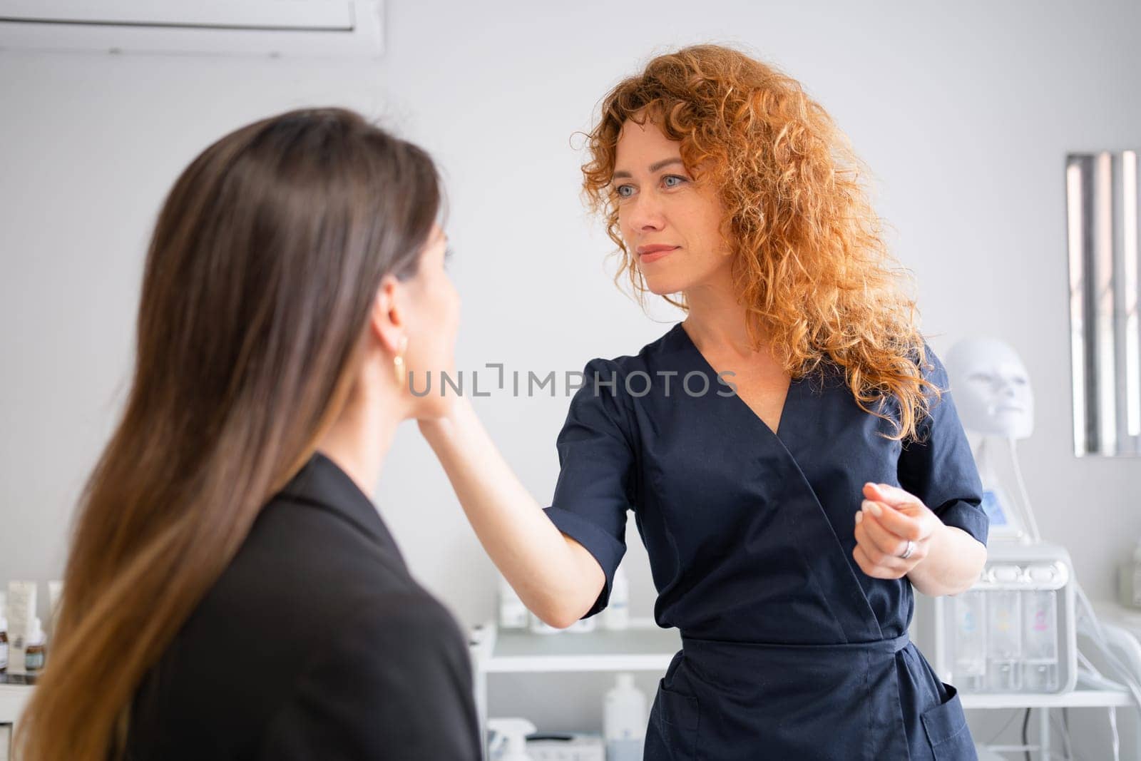 Woman at appointment at aesthetic clinic. Dermatologist consults patient about skincare. Female redhead professional beauty doctor touching face of her patient during medical check-up