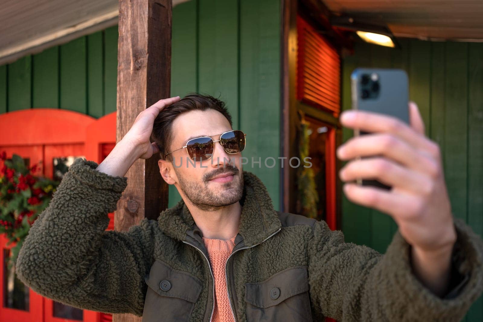 Man taking selfie outdoors. Stylish man wearing sunglasses making photo of himself with cell phone outside wooden house. Side view of handsome guy in fluffy jacket having video chat outdoor