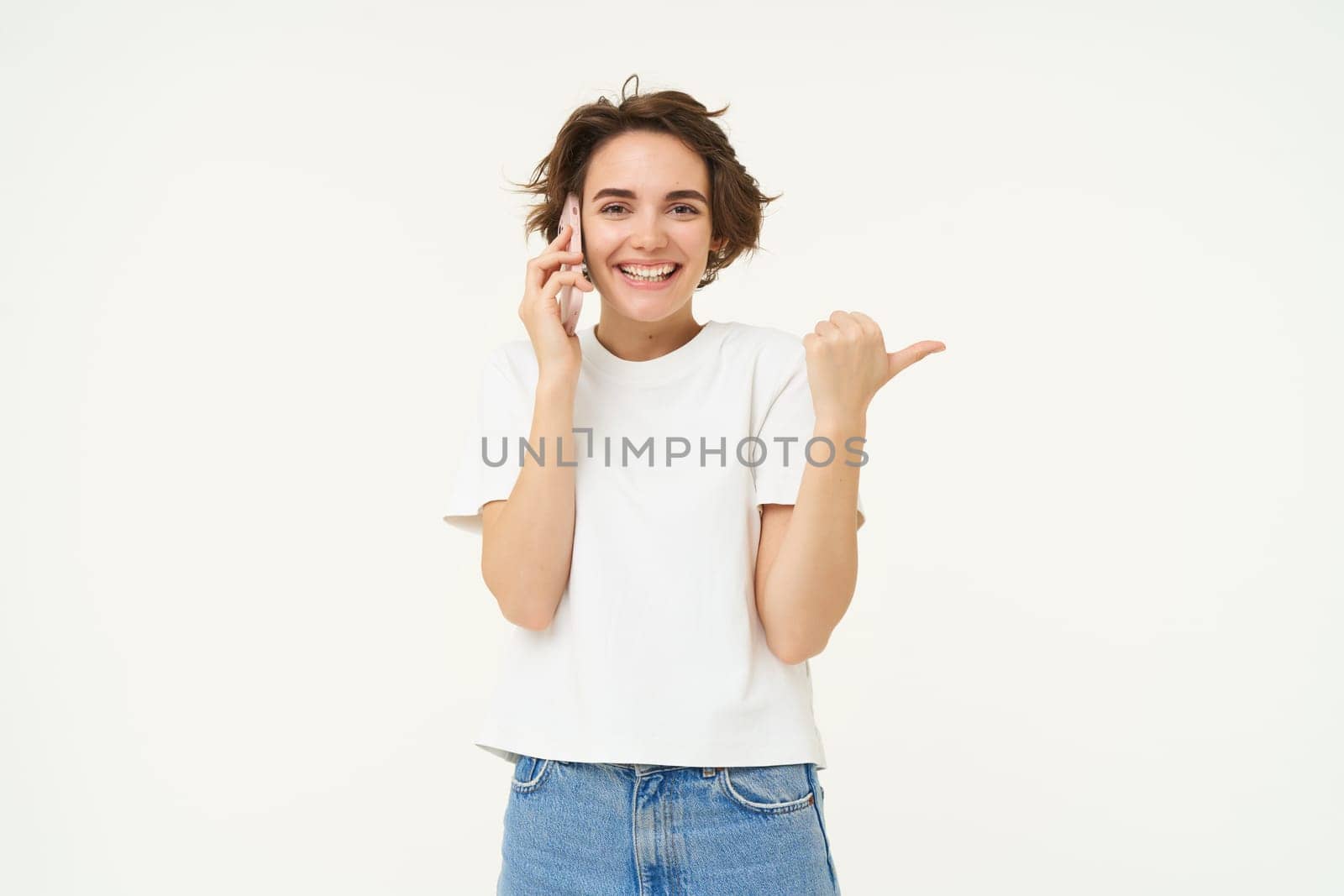 Portrait of friendly, smiling cute girl, pointing finger right, talking on mobile phone, showing smth aside on white copy space, isolated on studio background by Benzoix