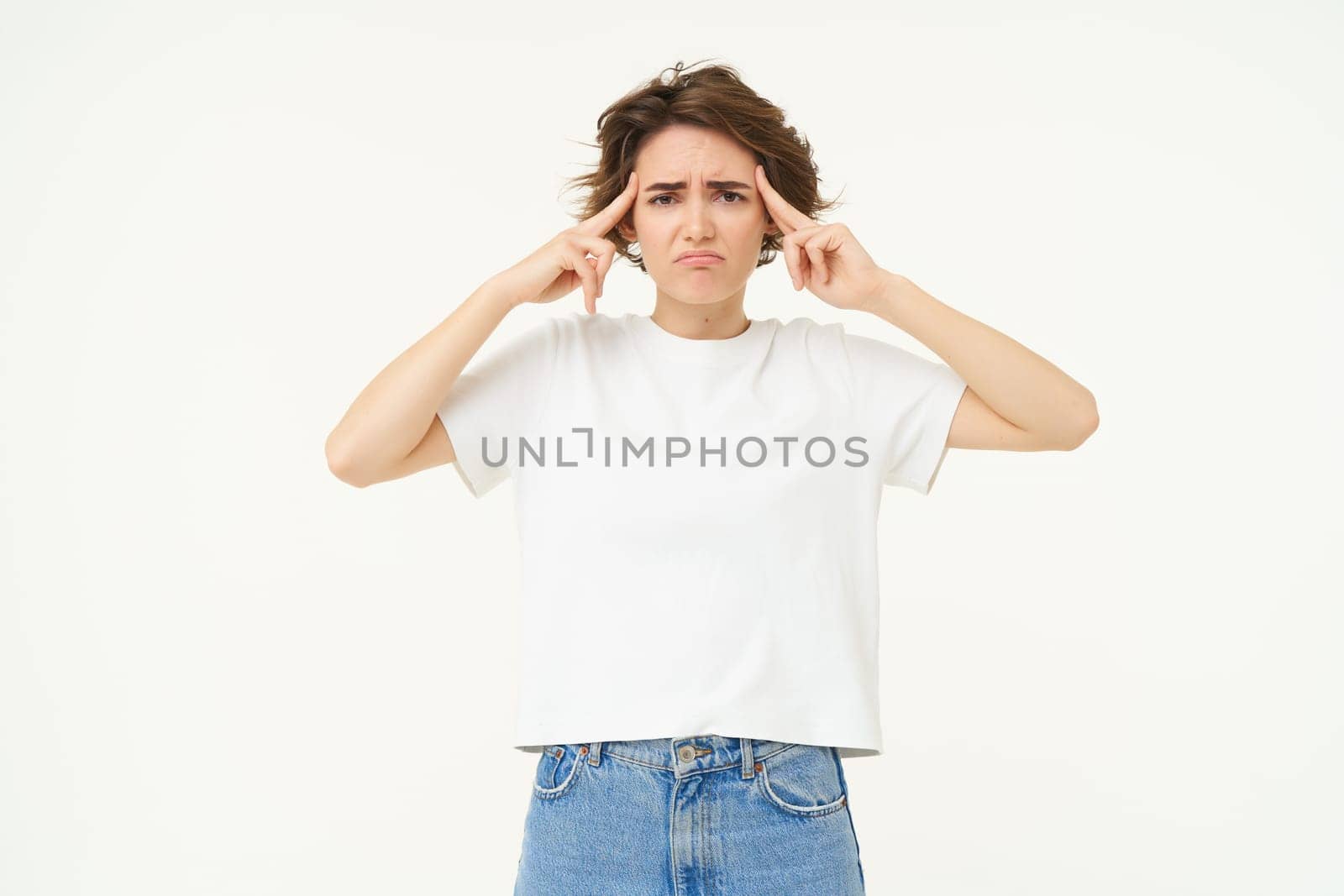 Image of brunette woman touches her head and frowns, looks anxious, thinking, trying to remember, looking worried and concerned, standing over white background.