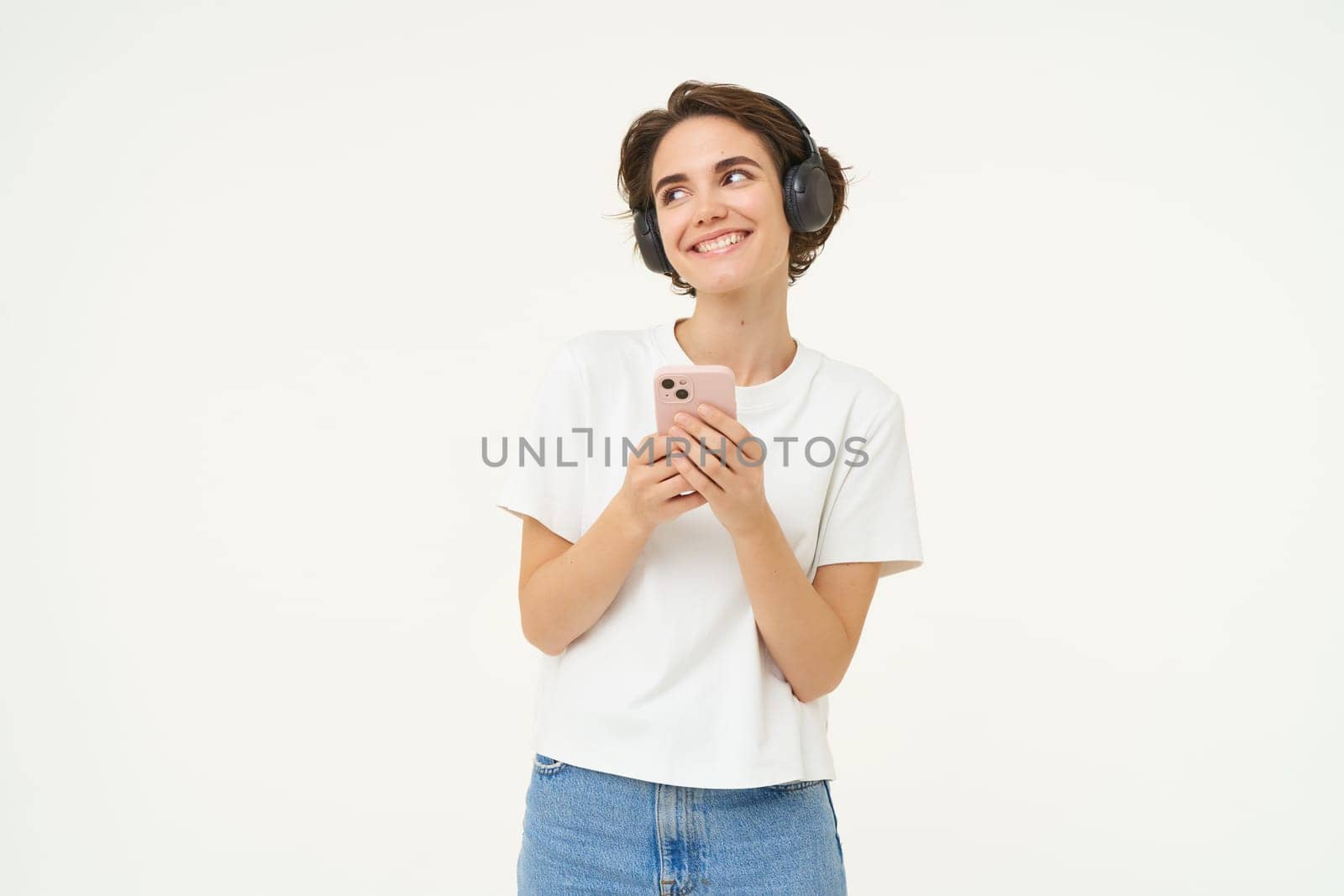 Image of happy young girl listens to music in headphones, standing with smartphone, using mobile phone streaming app, white background.