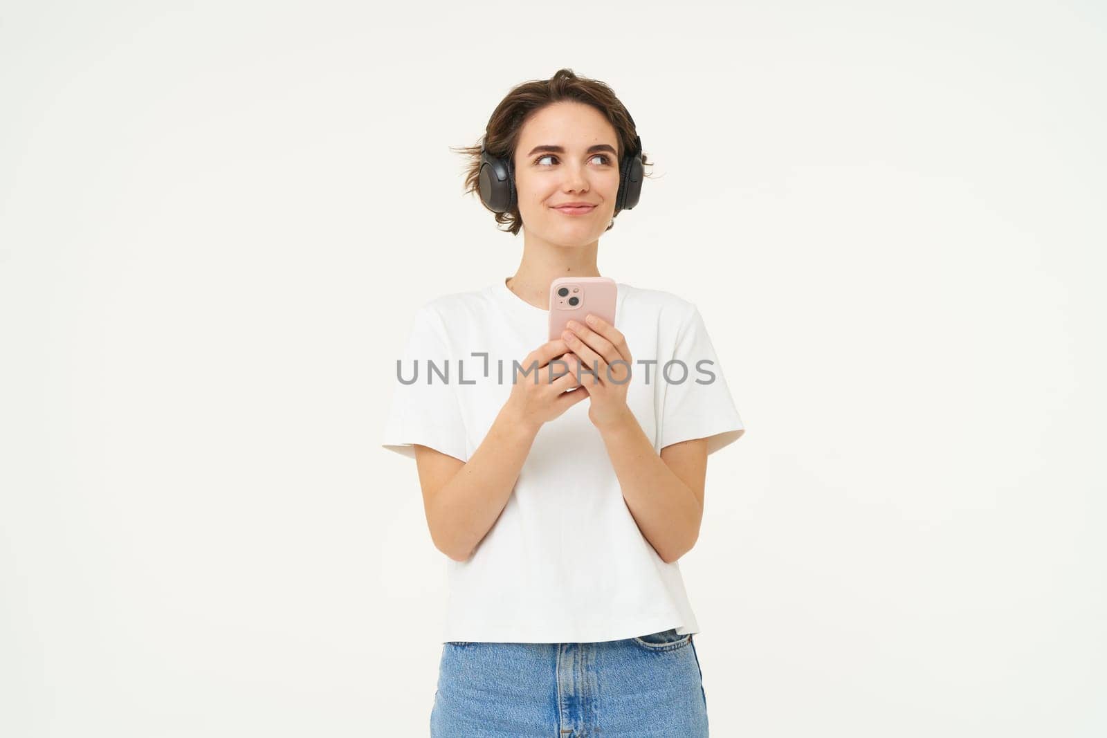 Portrait of smiling brunette woman with smartphone, smiling, looking aside, listening to music, standing over white studio background.