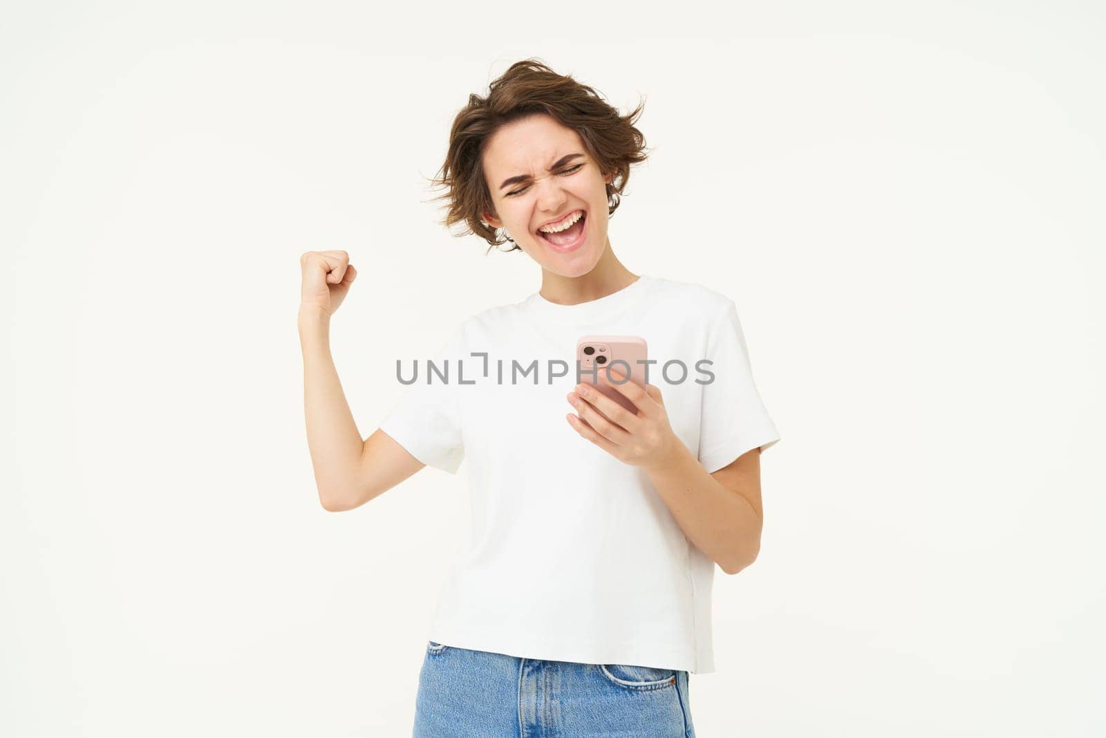 Portrait of excited brunette woman, holding smartphone and celebrating big win, achieves goal in mobile app, stands over white background. Copy space