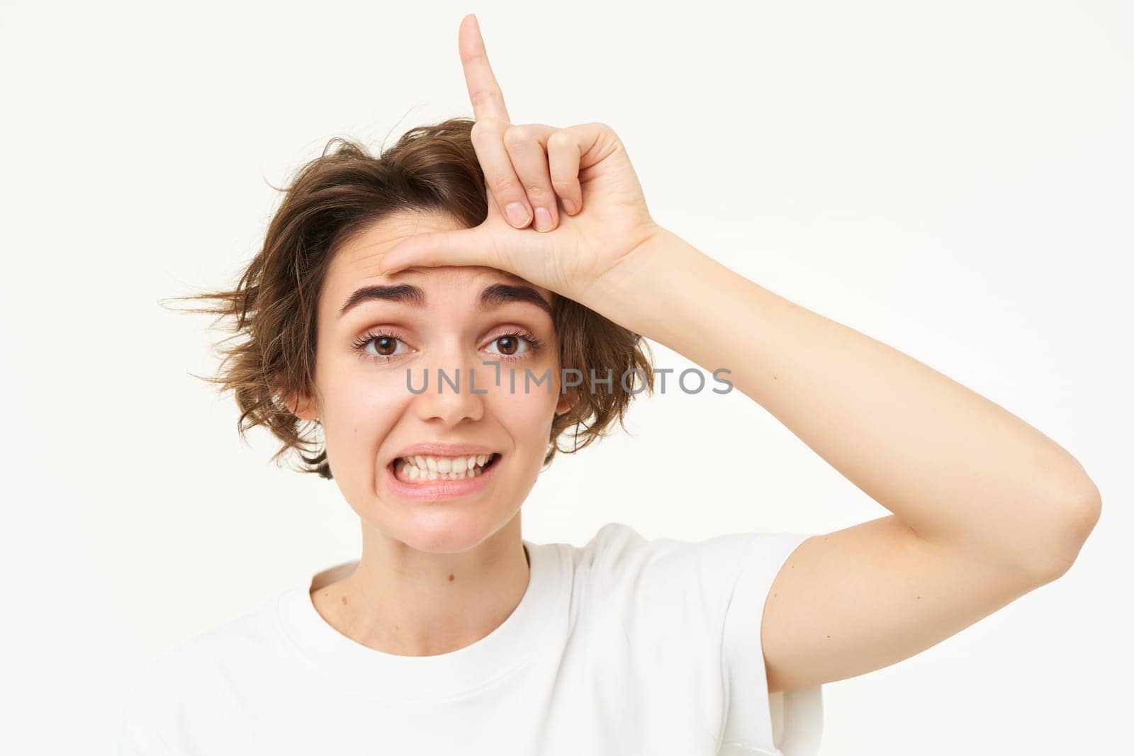 Close up of young woman making fun of friend, smiling and showing loser gesture, l letter on forehead, standing over white background.